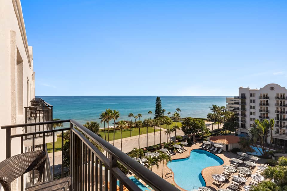 Beachfront hotel balcony with ocean view, pool deck below, palm trees, and sandy beach