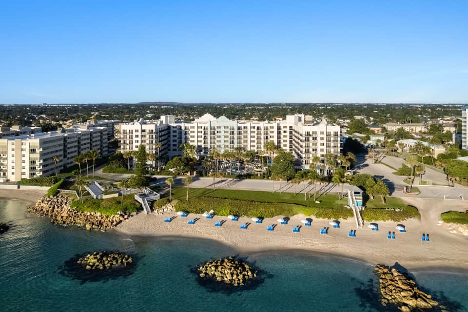 Beachfront resort with multi-story buildings, sand, blue umbrellas, and ocean view