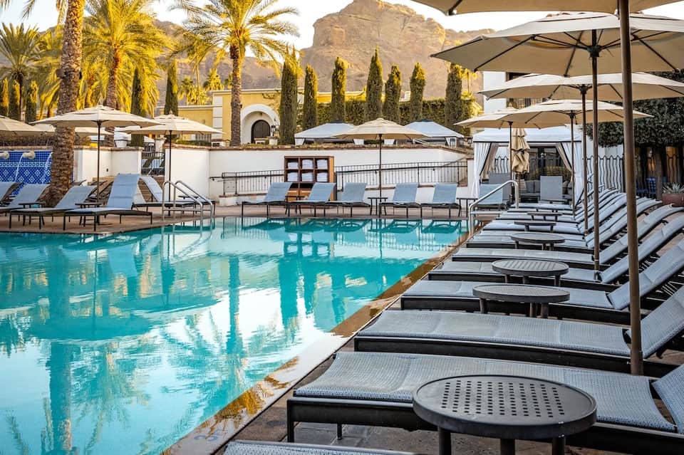 Resort pool with turquoise water, black lounge chairs, tan umbrellas, mountains, and desert landscaping