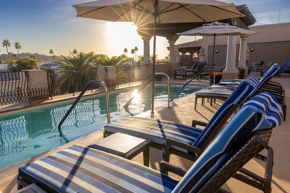 Rooftop pool deck with blue lounge chairs, umbrellas, mountain views, and desert landscape at sunset
