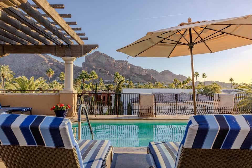 Resort pool with mountain views, umbrellas, striped lounge chairs, and pergola shade structure