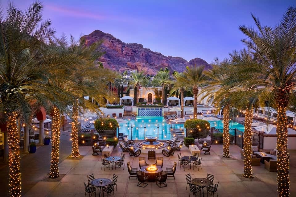Luxury resort pool deck at twilight with lit palm trees, fire pits, lounge seating, and mountain backdrop