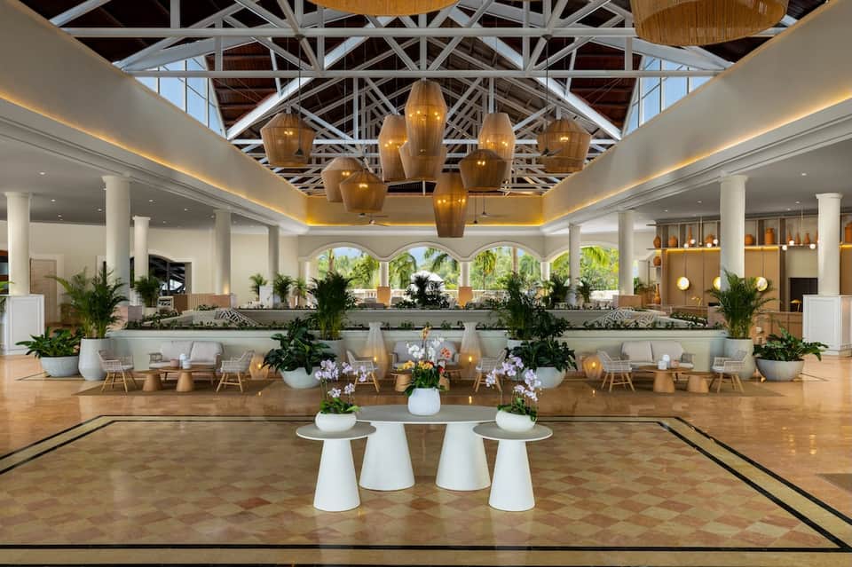 Grand hotel lobby with soaring white ceilings, brass pendant lights, white columns, tropical plants, and elegant seating