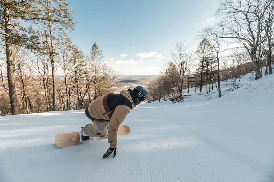 Snowboarder carving down snowy mountain slope with forested hillside and valley view in background