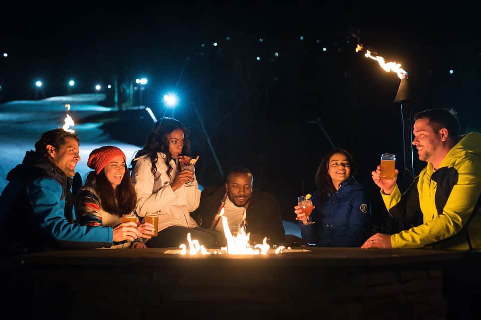 Group of friends sitting around a fire pit by the pool at night celebrating with drinks
