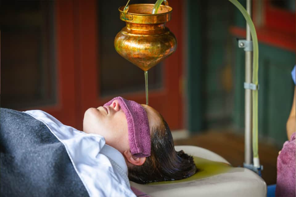 Spa treatment with oil pouring from brass vessel above woman's face during wellness service