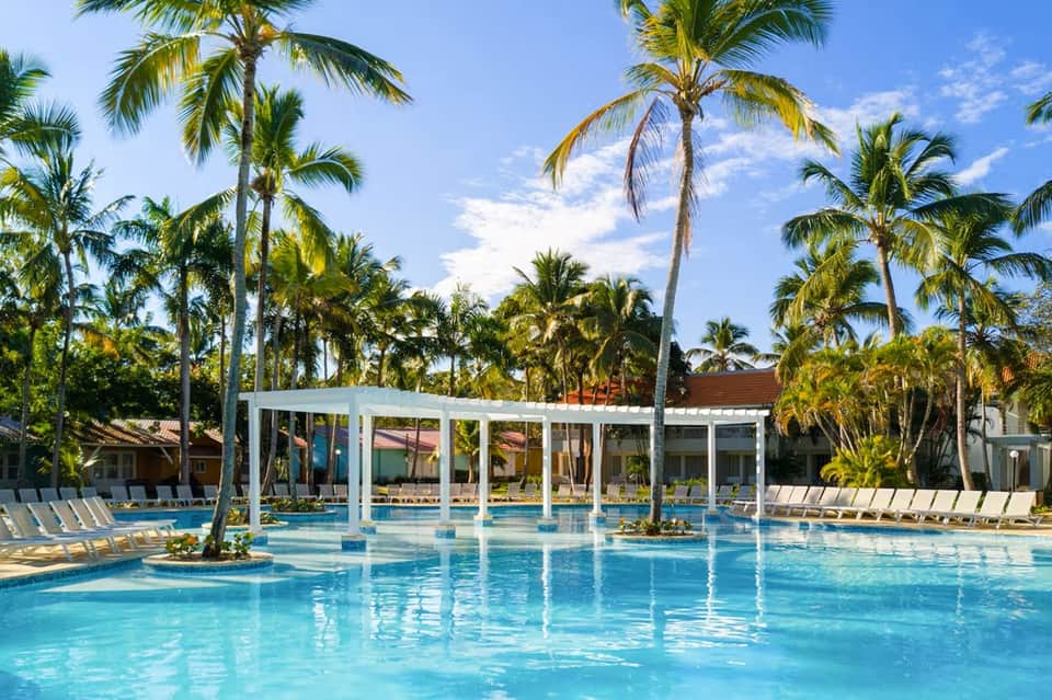 Resort swimming pool with pergola, lounge chairs, and tropical palm trees
