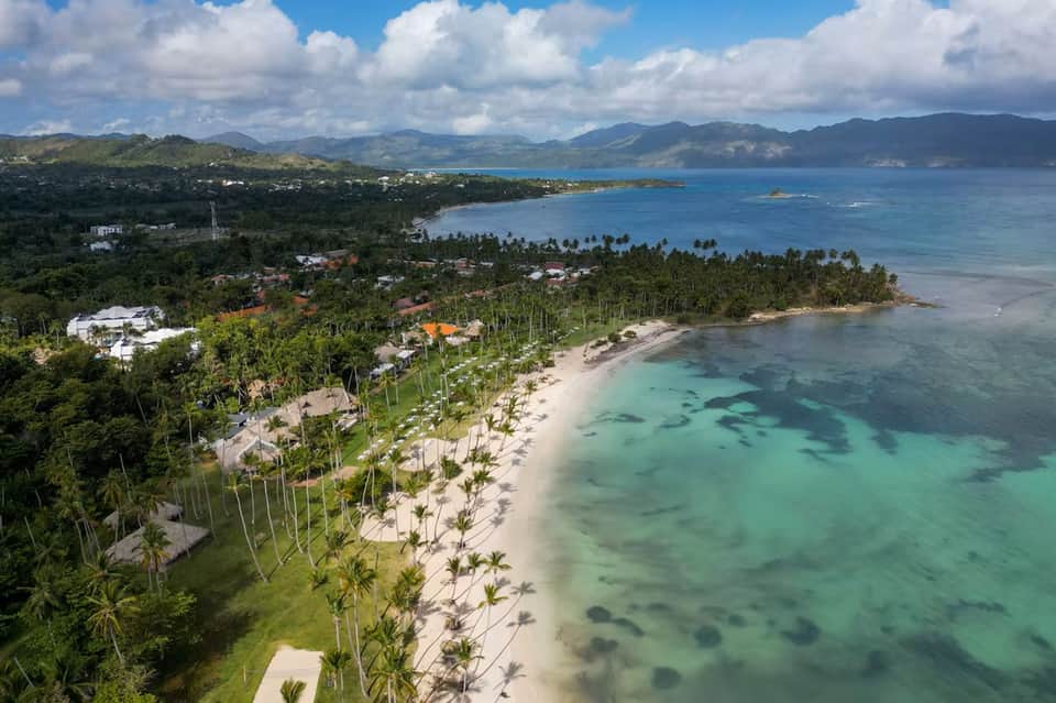 Aerial beach resort view with white buildings, palm-lined property, turquoise water, and mountains