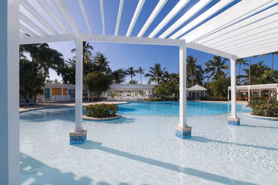 Luxury resort pool with white pergola, shallow wading area, palm landscaping, and modern white cabanas