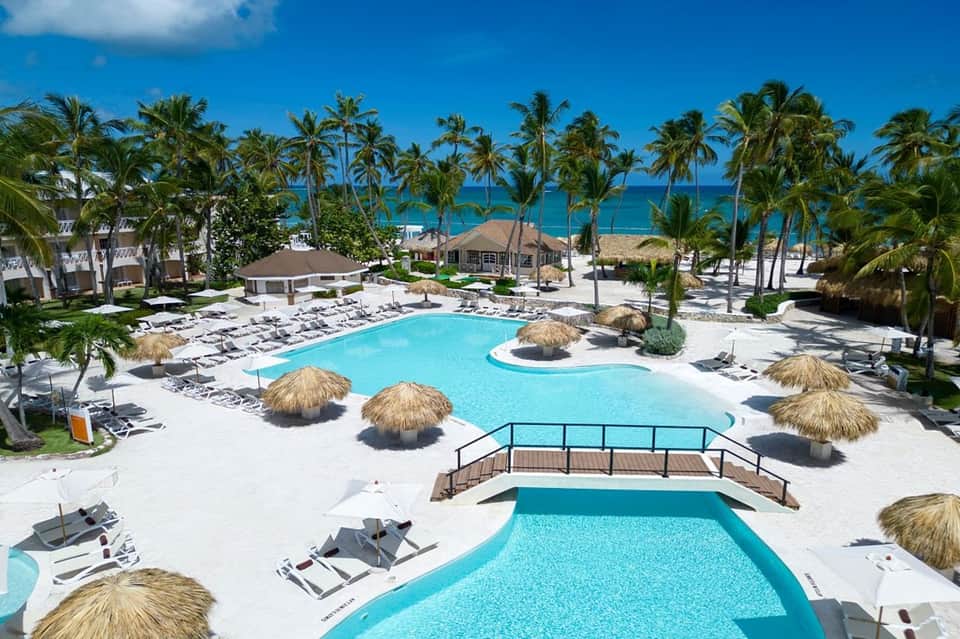 Beachfront resort pool complex with turquoise water, palm trees, tiki umbrellas, and ocean view