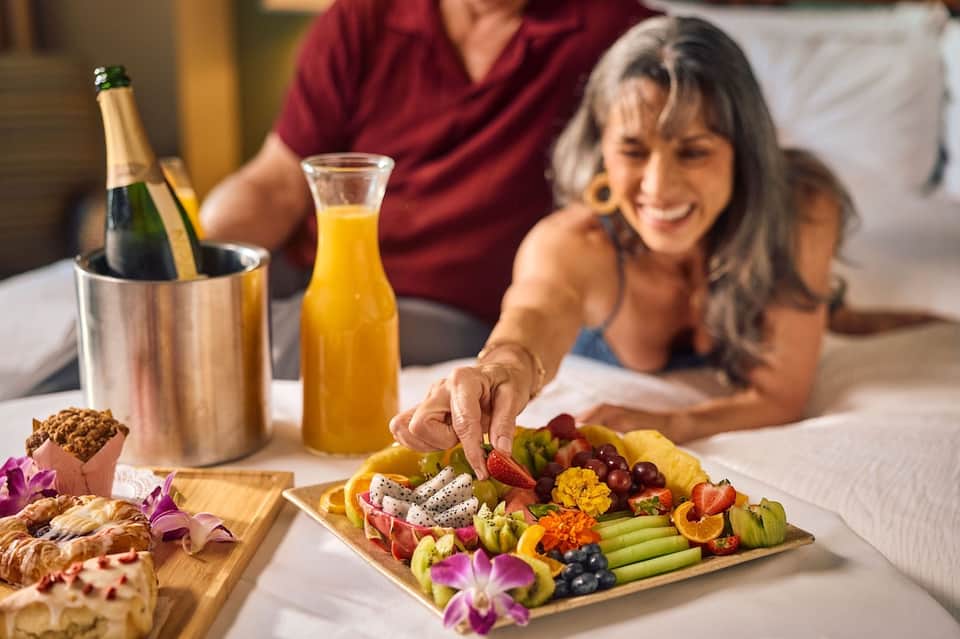 Couple enjoying in-room amenities with champagne, fruit platter, and sparkling cider on bed