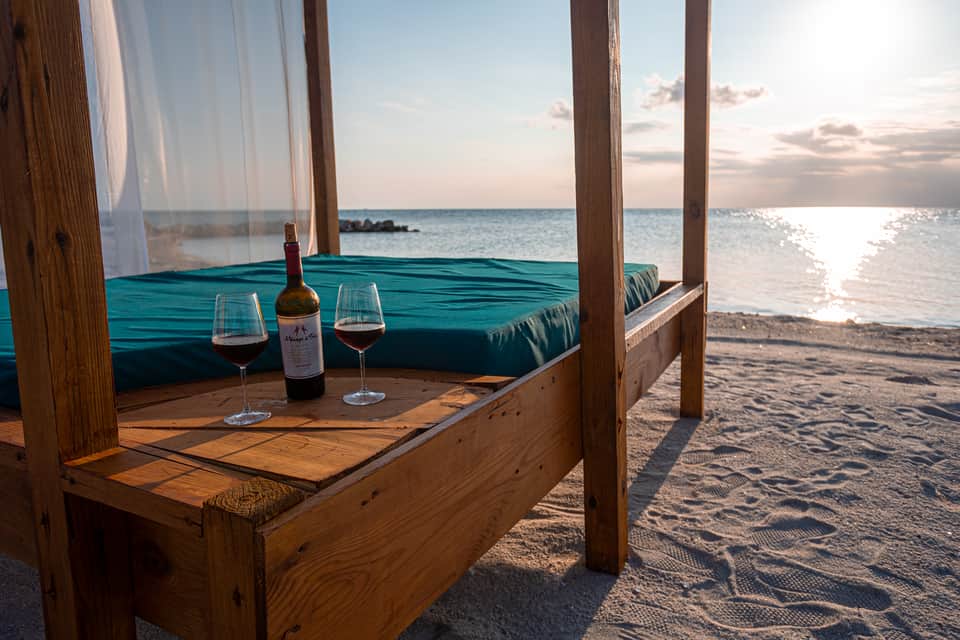 Beachfront cabana with turquoise cushions, wine glasses, and ocean sunset view with sheer curtains