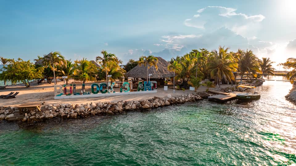 Beach village with colorful sign, palm trees, thatched structures, and turquoise water