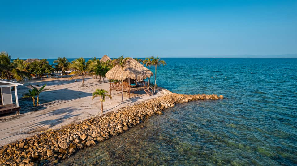 Tropical beachfront resort with thatched-roof structures, palm trees, sandy beach, and calm turquoise water