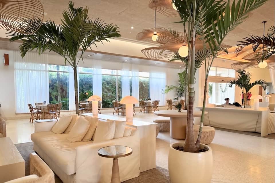 Bright hotel lobby with cream seating, tall palm plants, pendant lights, and glass doors