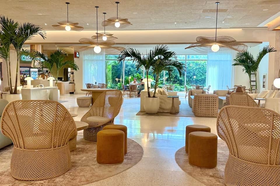 Modern hotel lobby with natural lighting, woven chairs, palm plants, and café area