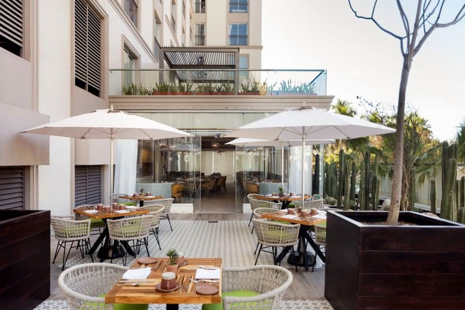 Modern outdoor dining terrace with tables, large umbrellas, glass walls, and tropical landscaping