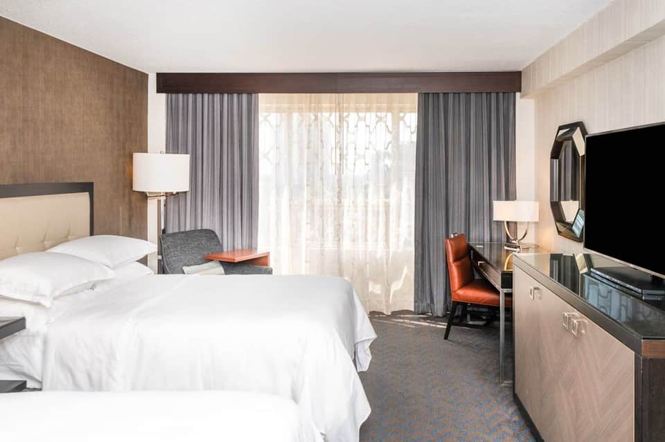 Hotel bedroom with queen bed, modern furnishings, work desk, balcony doors with curtains, and flat-screen TV