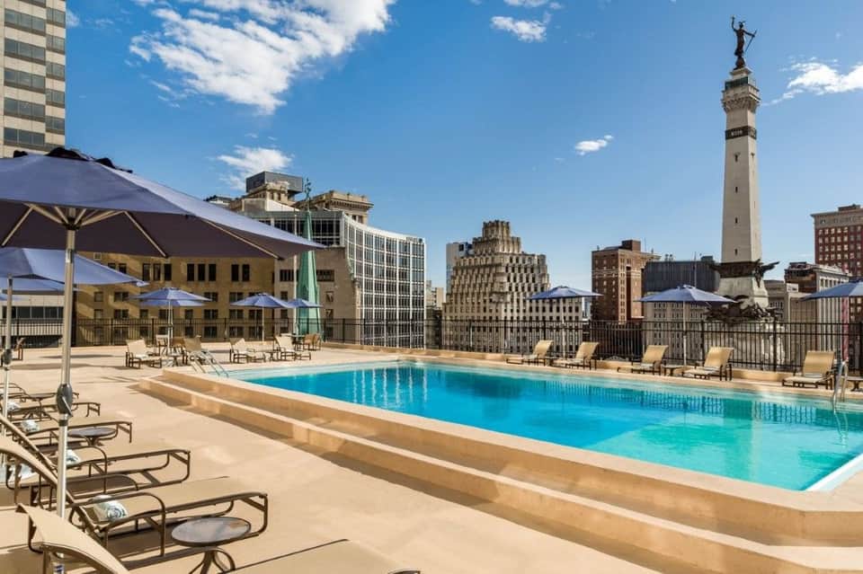Rooftop pool in urban setting with city skyline, blue umbrellas, and lounge chairs
