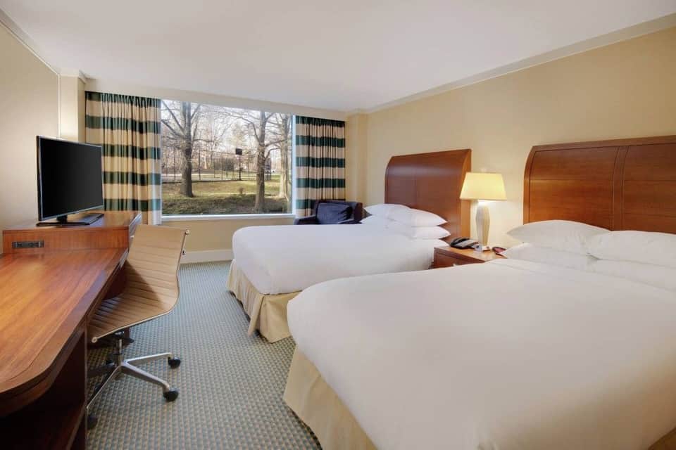 Hotel bedroom with two queen beds, wood headboards, TV, and large window with park views