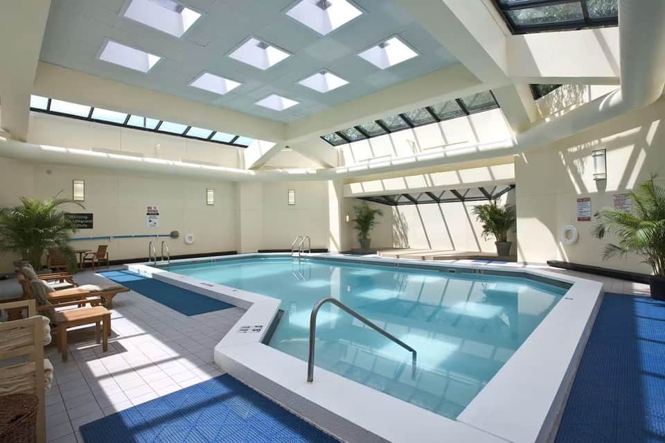Bright indoor pool facility with skylights, spa tub, lounge seating, and tropical plants