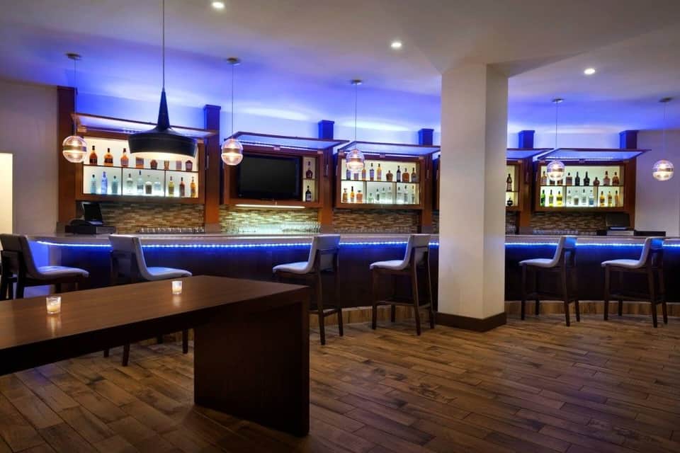 Modern hotel bar with blue ambient lighting, multiple bar stations, and dark wood furnishings