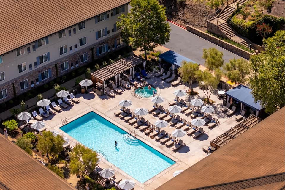 Aerial view of resort pool with lounge chairs, umbrellas, hot tub, and landscaping