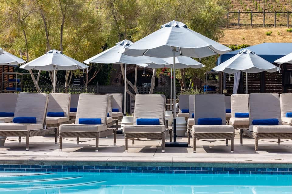 Resort pool area with white umbrellas, lounge chairs with blue cushions, and hillside setting