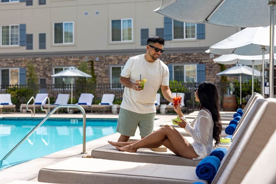 Couple enjoying beverages poolside under umbrellas at resort with building and landscaping backdrop