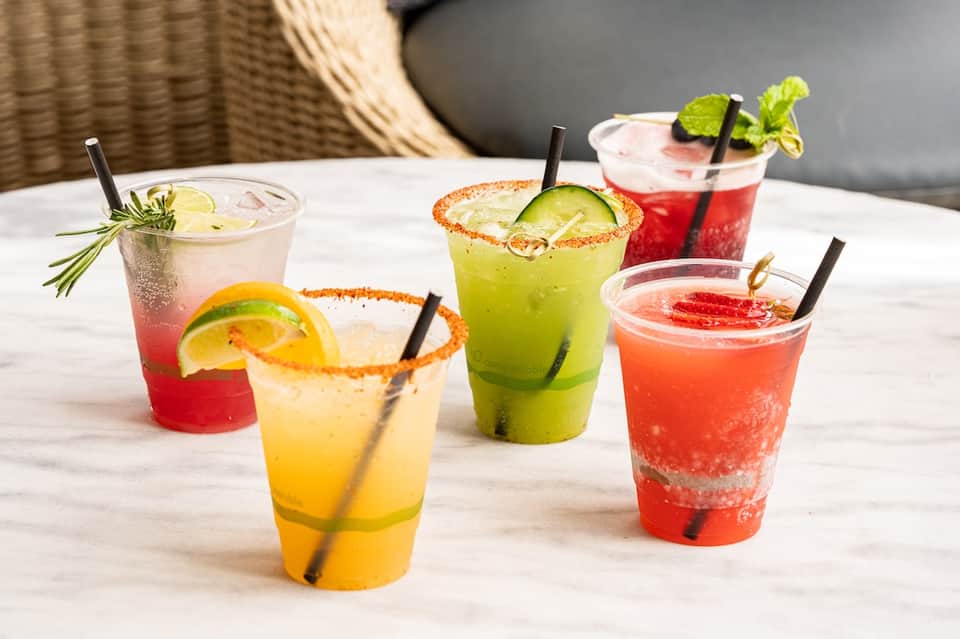 Colorful cocktails displayed on white table with garnishes including citrus slices, cucumber, and fresh herbs