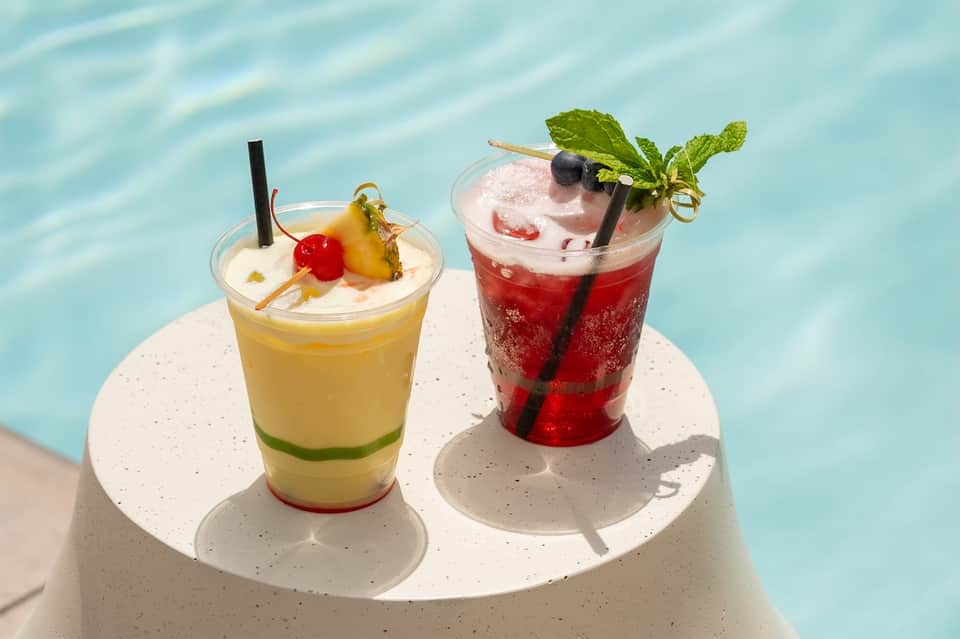 Two colorful cocktails on white table by turquoise pool water with garnishes and ice