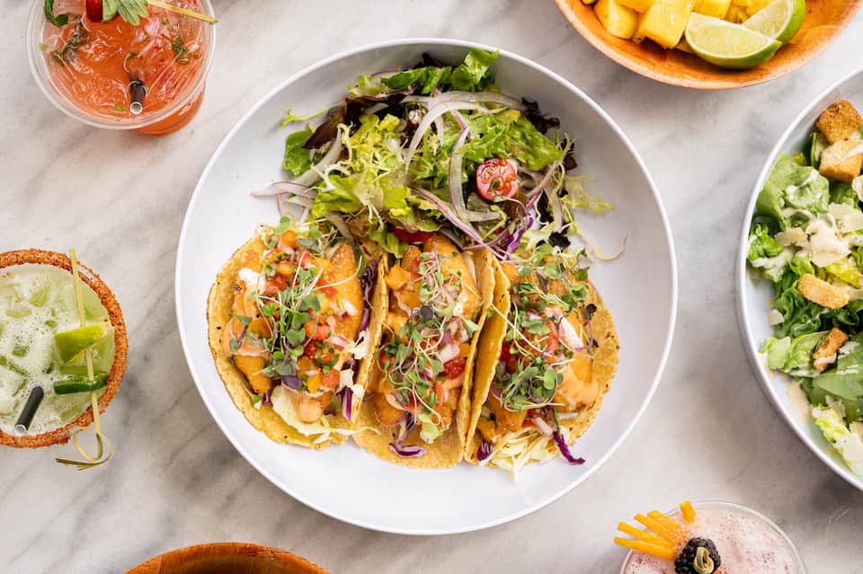 Overhead view of colorful tacos with fresh salad, lime, and tropical cocktails on table