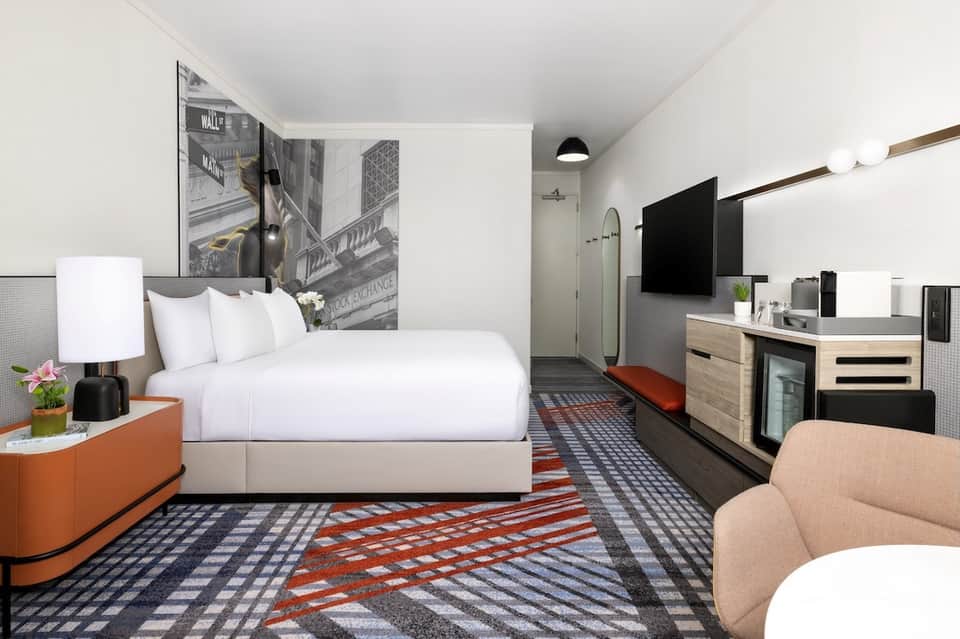 Modern hotel room with white bedding, orange accents, geometric patterned rug, TV, and contemporary furnishings