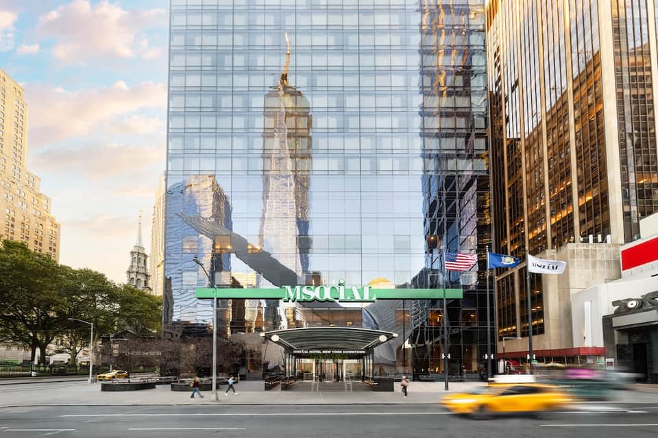Urban hotel entrance with modern glass facade reflecting historic buildings, green signage, and city street traffic