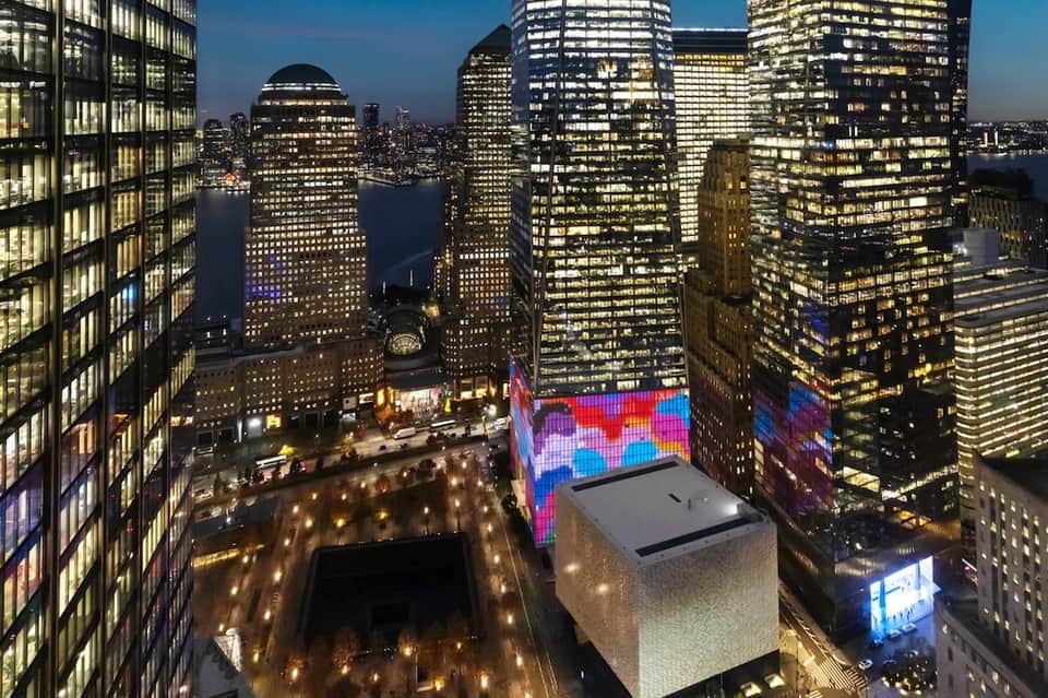 Nighttime aerial view of illuminated skyscrapers surrounding plaza with digital billboard and water feature