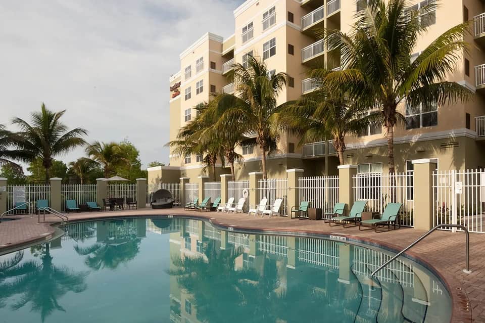Resort pool area with loungers, white fencing, multi-story hotel building, and palm trees