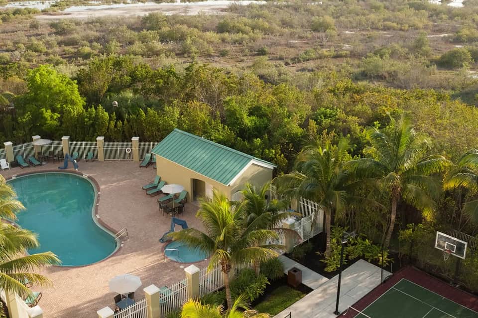 Aerial view of resort pool surrounded by palm trees, white fencing, cabana structure, sports court, and forested landscape
