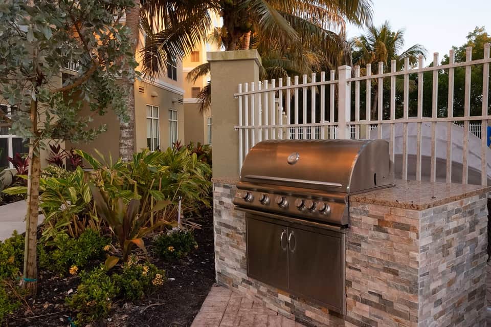 Backyard grilling area with stainless steel barbecue, brick surround, white fence, and tropical landscaping