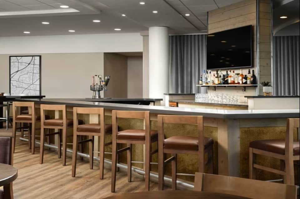 Modern hotel bar and lounge area with wooden bar stools, shelving, and contemporary furnishings