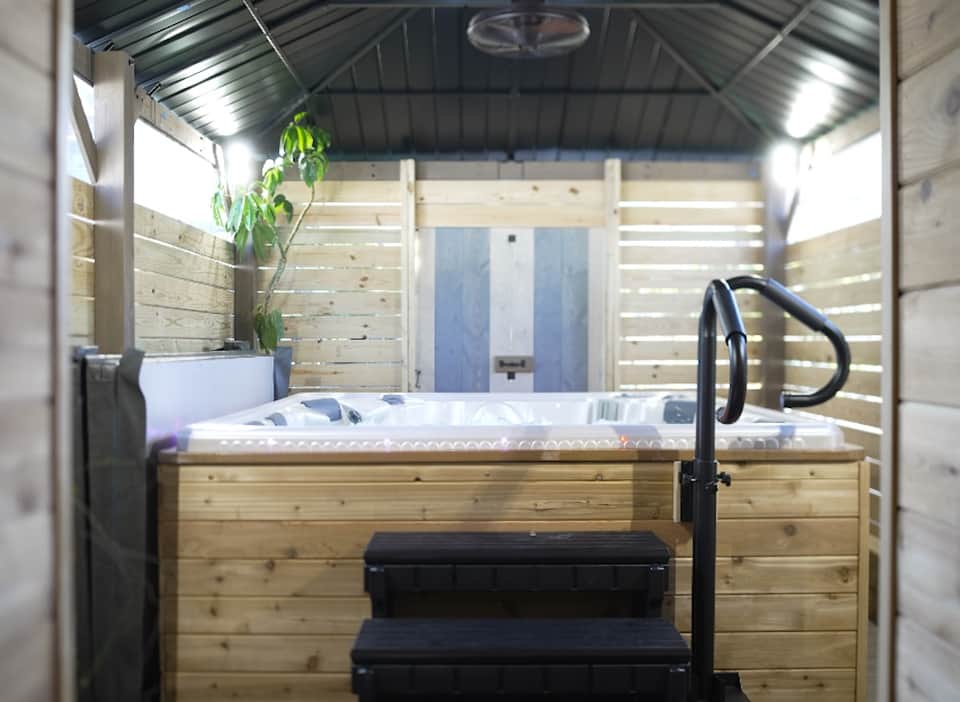 Wooden hot tub with black spa steps and railings inside covered wooden structure with natural light