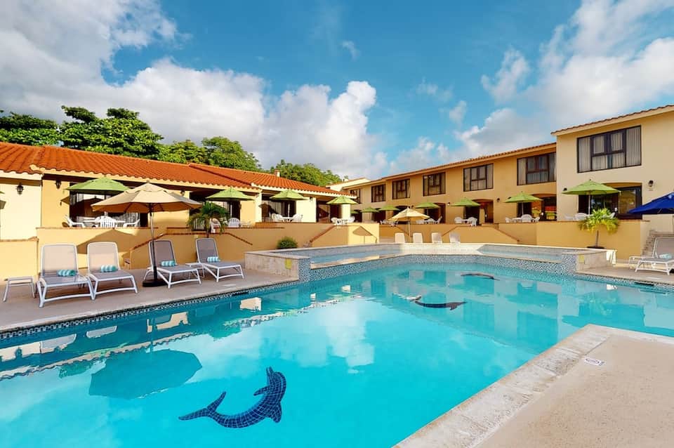 Tropical resort pool with mosaic tile design, loungers, yellow umbrellas, and Spanish-style architecture