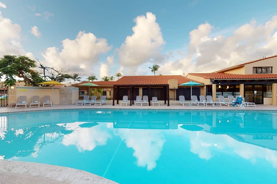 Resort pool reflecting sky with cabanas, lounge chairs, palm trees, and Spanish-style buildings