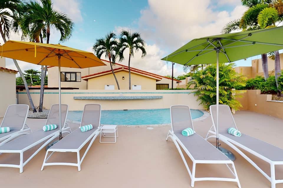 Resort pool with white lounge chairs, colorful umbrellas, palm trees, and tropical building architecture