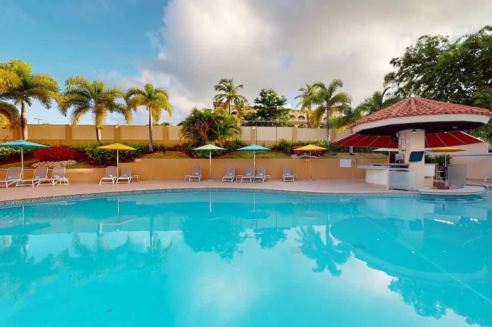 Resort swimming pool with turquoise water, red umbrellas, lounge chairs, and tropical vegetation