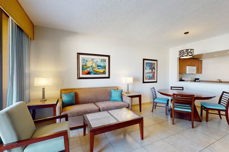 Hotel suite living area with sofa, dining table, kitchenette, and tropical-themed artwork