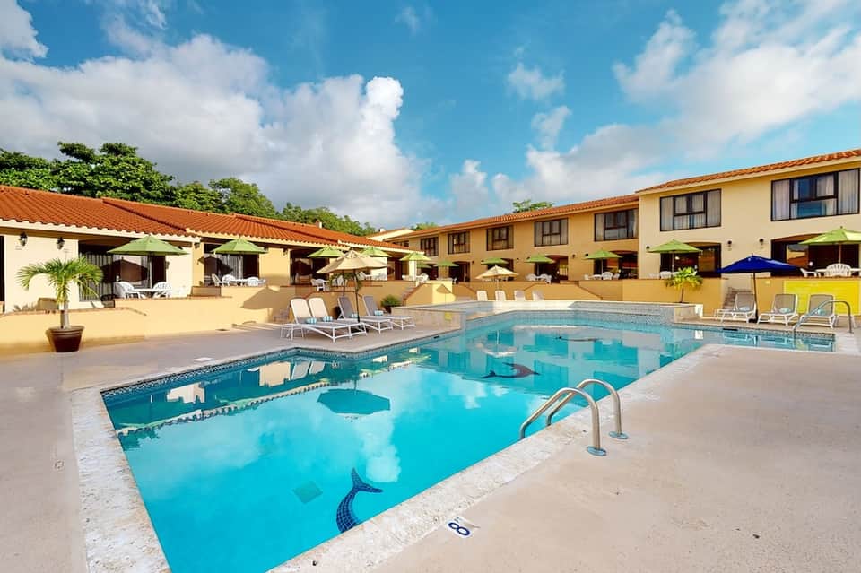 Turquoise pool with lap lanes, lounge chairs, umbrellas, and tan Spanish-style buildings