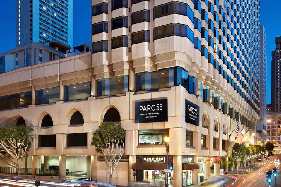 Historic sandstone hotel building facade with arched windows, modern signage, street-level retail, and urban surroundings.