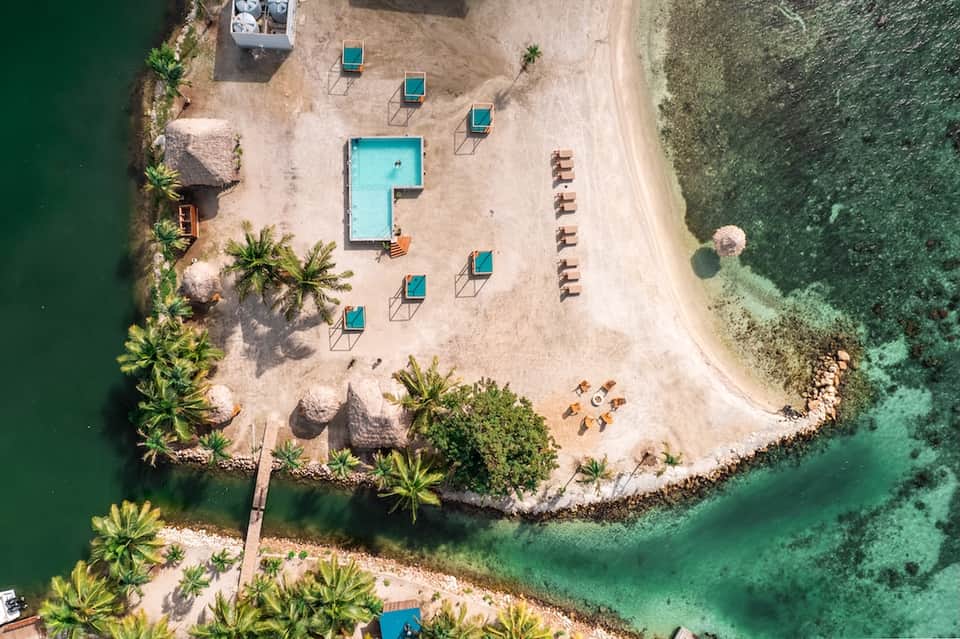 Aerial beach view with turquoise pool, lounge chairs, palm trees, and clear green ocean water