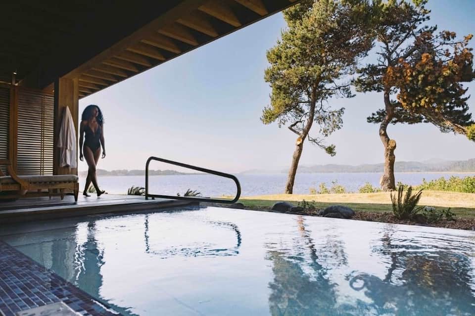 Woman in swimwear approaching natural hot spring pool overlooking lake and forested landscape