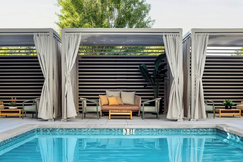 Modern poolside cabana with sheer curtains, lounge seating, and swimming pool reflection
